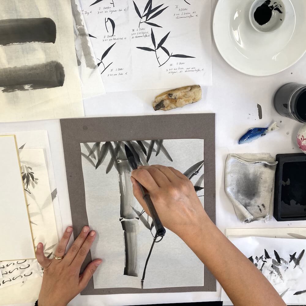 Hand mit Sumi-e-Pinsel beim Malen eines Bambusmotivs in schwarzer Tusche auf Papier.